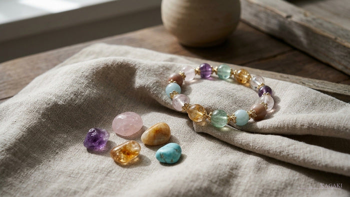 Multi-stone KAGAKI birthstone bracelet with rose quartz, amethyst, citrine, amazonite, fluorite and aquamarine beads beside loose tumbled birthstones on cream linen, editorial hero image for a birth month bracelet guide.