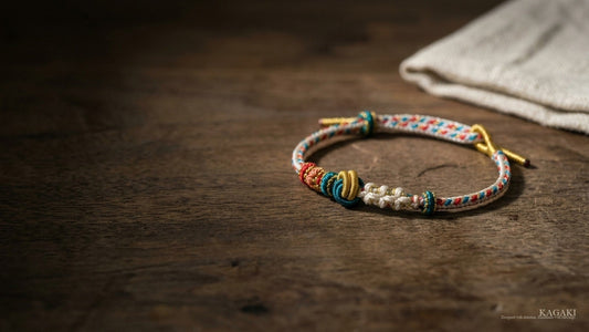 KAGAKI Spectrum handmade multi-color Tibetan knot bracelet at rest on aged walnut beside a single folded linen in soft afternoon daylight, shown as an editorial hero image for a guide on bracelet color meaning in feng shui.