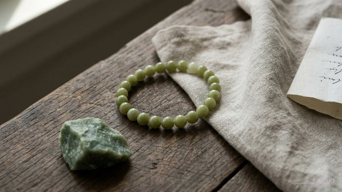 Handmade KAGAKI natural green jade beaded bracelet at gentle rest on aged wood beside a raw nephrite jade stone and a folded calligraphy note — a quiet editorial image for a guide to jade bracelet meaning and symbolism.