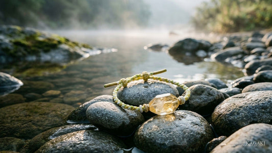 A raw citrine bracelet on knotted cord resting on wet river stones at dawn, mist low over the stream.