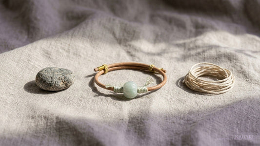 A jade barrel bead on leather cord laid on linen between a rough grey stone and a small coil of undyed cord.