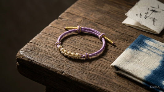 KAGAKI Japanese Zen Sakura handmade cord bracelet at gentle rest on aged walnut beside a folded cream-and-indigo natural textile in soft afternoon daylight, shown as an editorial hero image for a guide on what a kumihimo bracelet is.