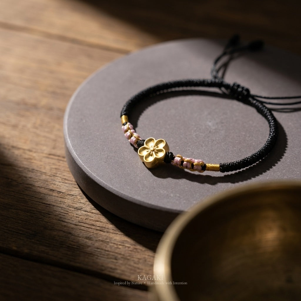 KAGAKI handmade cord bracelet sakura plum blossom cord bracelet (ink & petal) — black / green / red with gold accent, editorial styled scene.