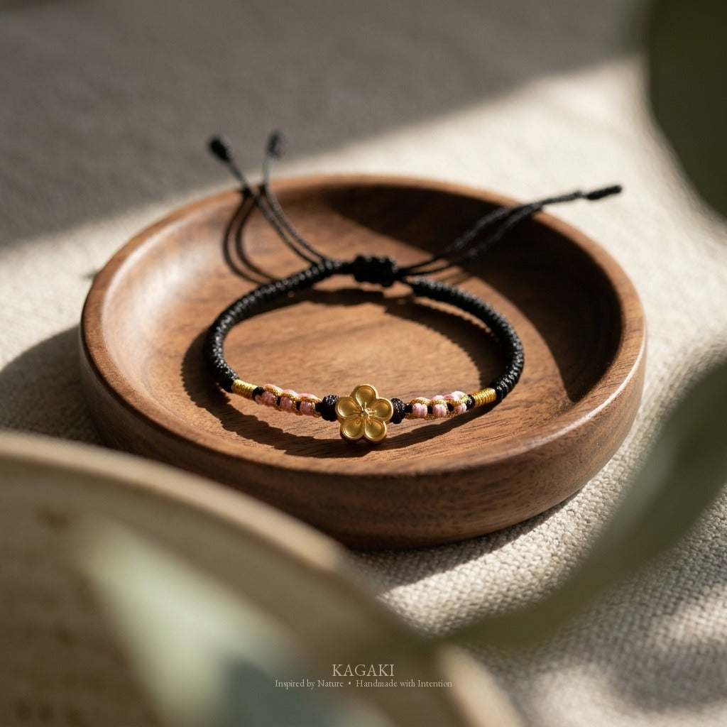 KAGAKI handmade cord bracelet sakura plum blossom cord bracelet (ink & petal) — black / green / red with gold accent, editorial styled scene.