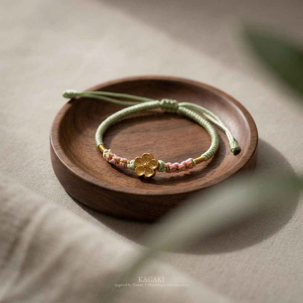 KAGAKI handmade cord bracelet sakura plum blossom cord bracelet (moss & petal) — black / green / red with gold accent, editorial styled scene.