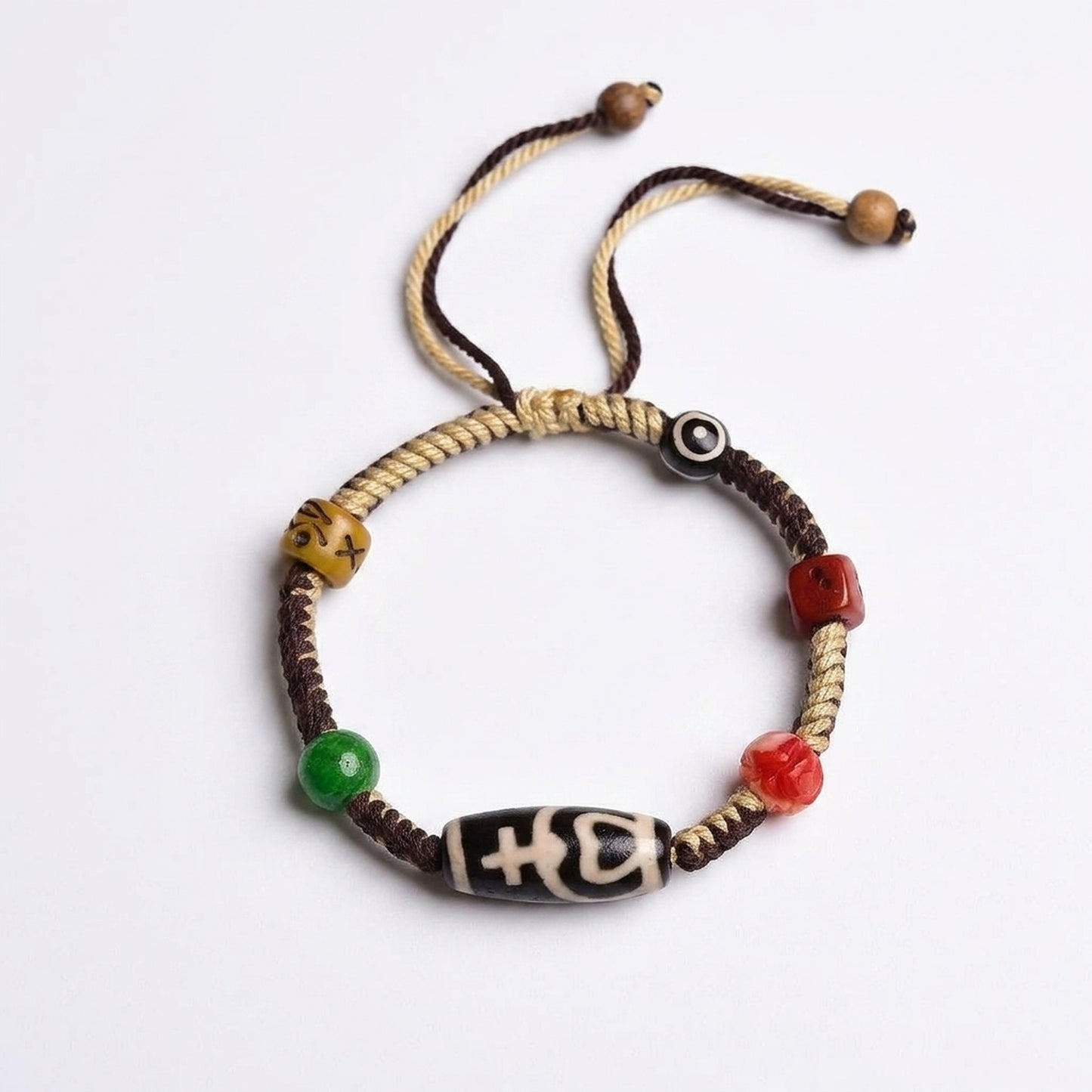 KAGAKI Dzi Tibetan Protection Bracelet — handmade two-tone braided cord bracelet with central black-and-white Dzi barrel bead, eye-Dzi, jade green, red carved, and ochre swirl beads, premium white-background product photograph.