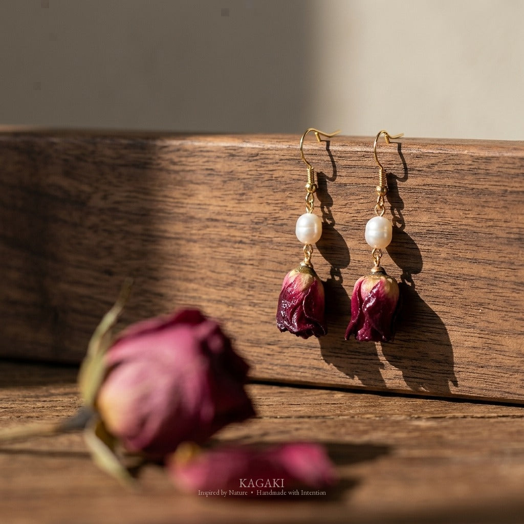 KAGAKI dried rose earrings styled on warm walnut with foreground dried rose petal blur, low angle showing wine-red rose color and freshwater pearl