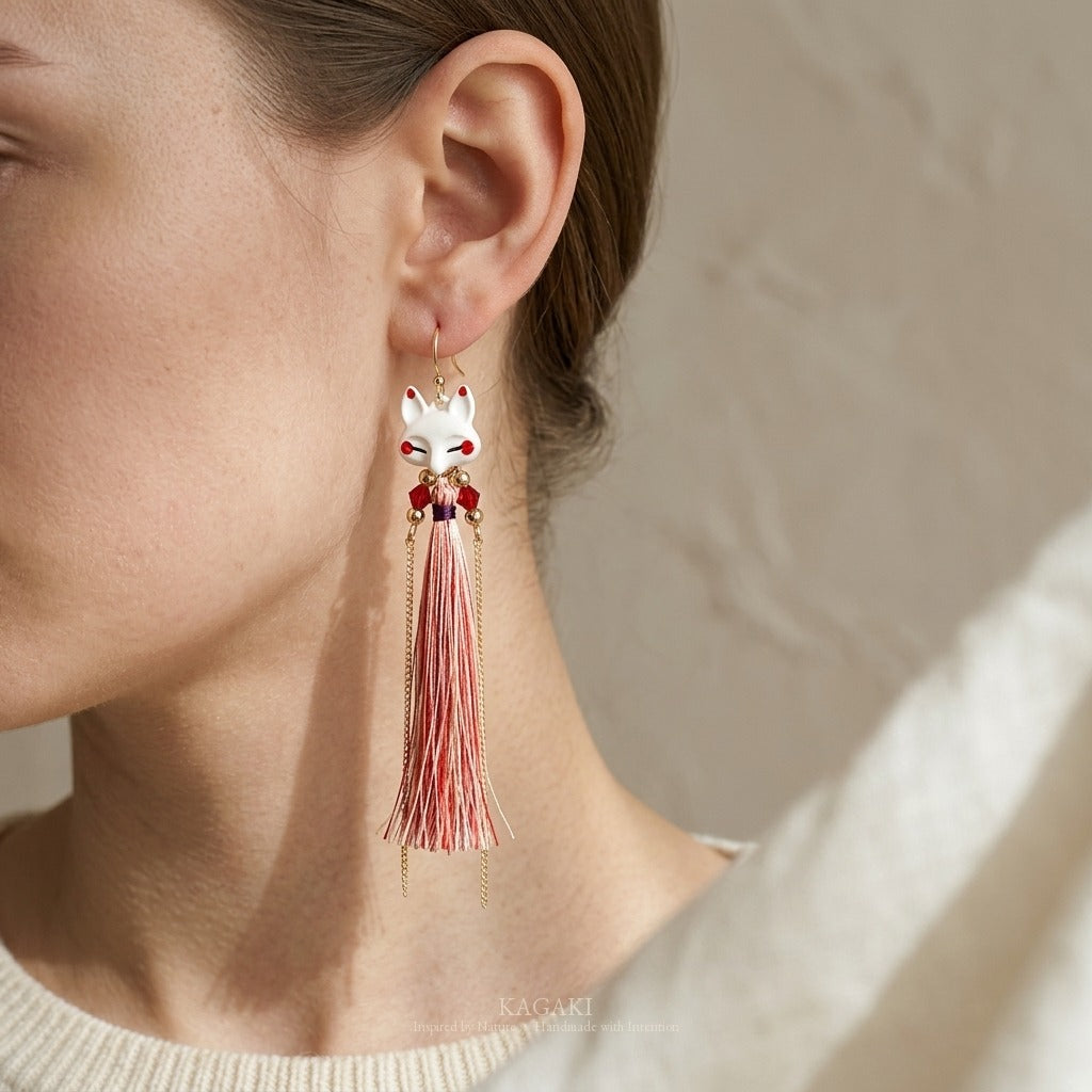 KAGAKI Japanese kitsune fox tassel earring shown worn on ear in side profile, the porcelain fox face and coral tassel falling along the neck