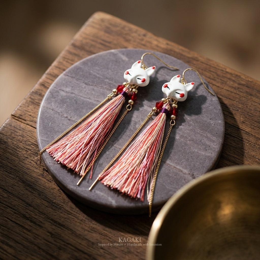 KAGAKI kitsune fox earrings styled on stone disc with brass bowl, showing the porcelain fox face and coral fine fiber tassel detail in editorial composition