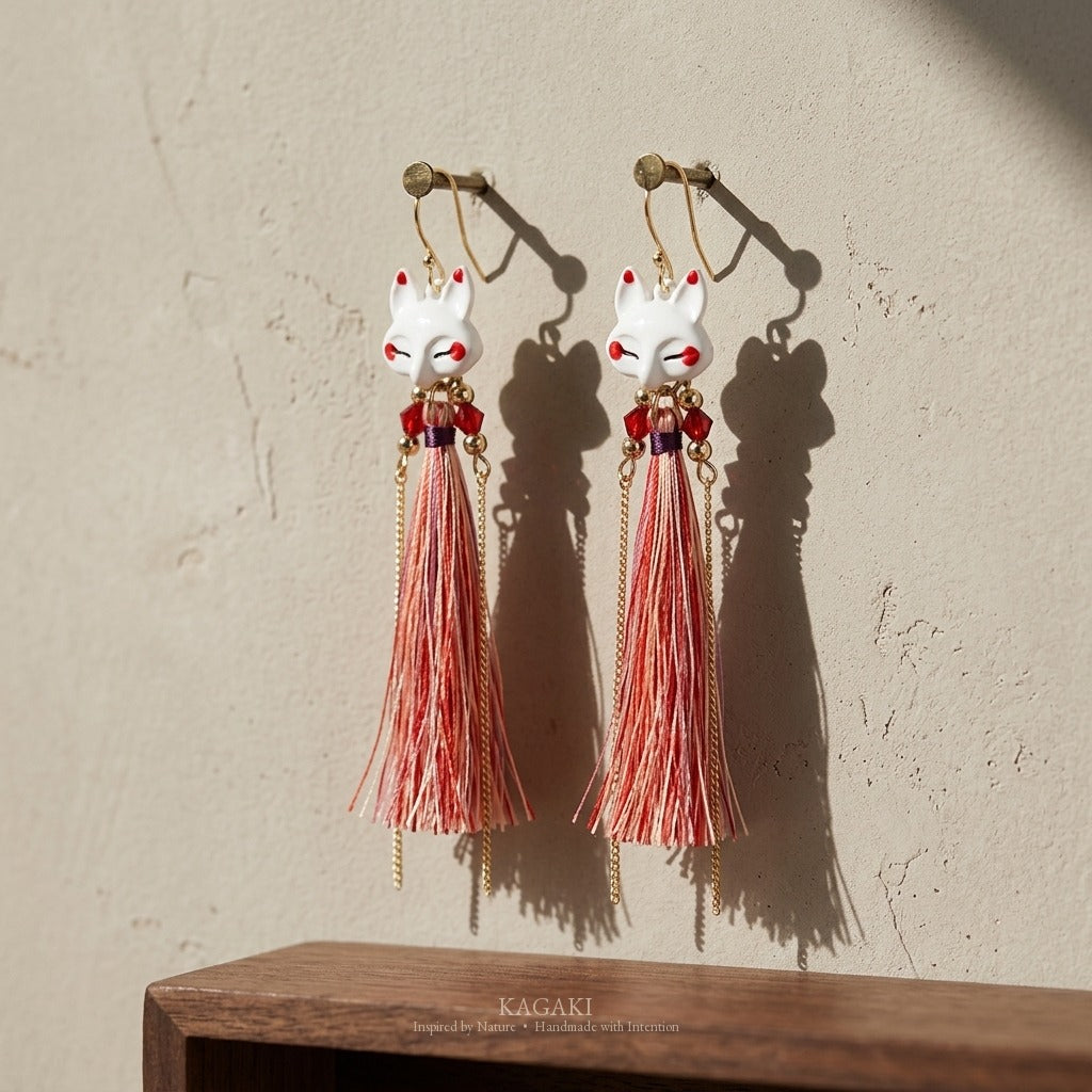 KAGAKI kitsune earrings hung on neutral plaster wall in vertical editorial composition, fox heads and fine fiber tassels casting soft architectural shadows