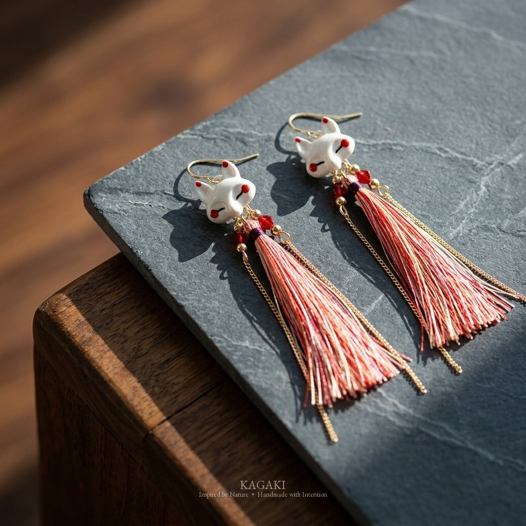 KAGAKI kitsune earring pair laid on dark slate over warm walnut, low angle showing the fox heads and cascading fine fiber tassels with their natural shadows