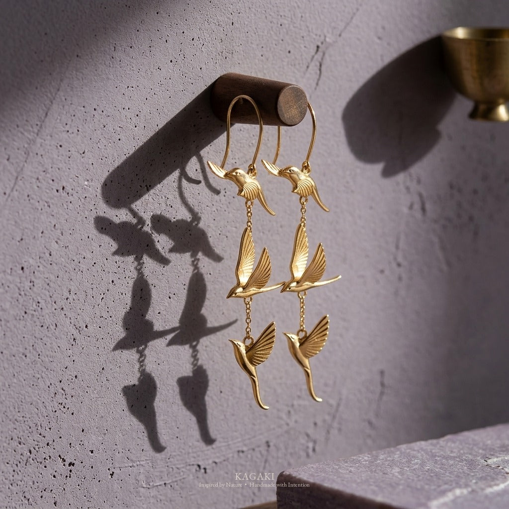 KAGAKI gold flying-swallow dangle earrings hanging from a dark walnut peg against smoky lavender plaster, with a sculptural cast shadow — quiet meditation altar editorial.