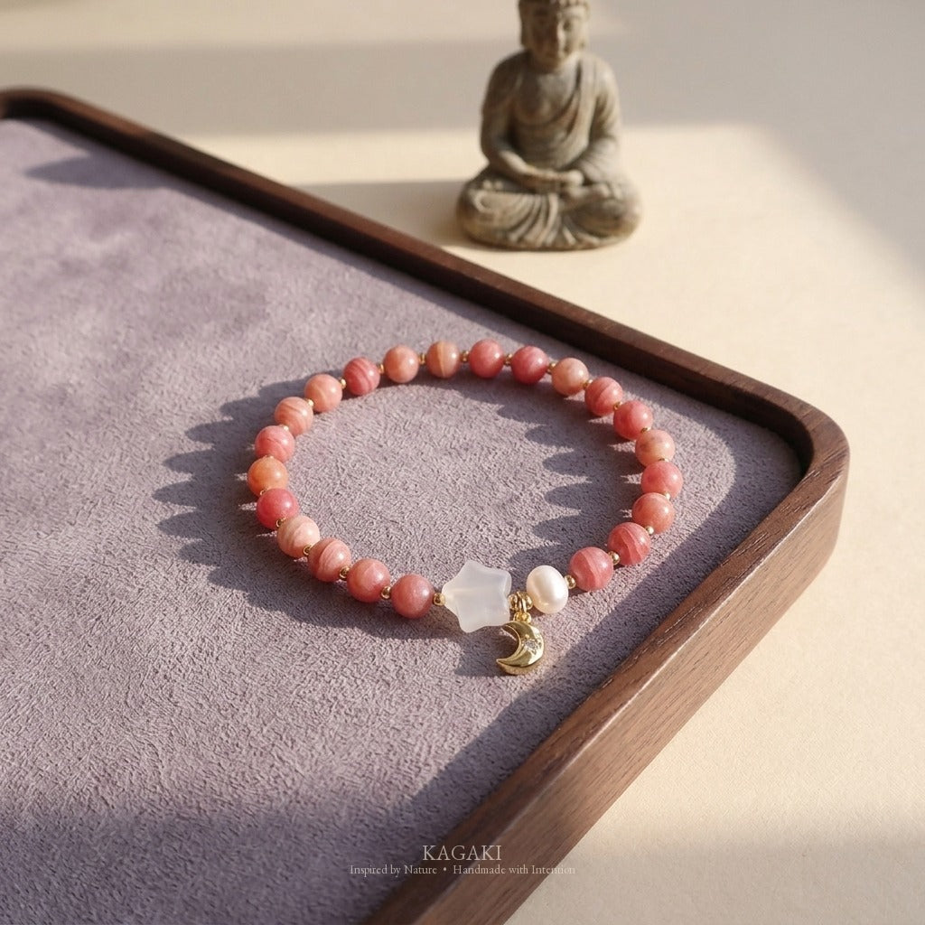 KAGAKI rhodochrosite bracelet with chalcedony star, pearl, and gold-tone crescent moon charm styled on a dark walnut tea tray with dusty-lavender suede inset and a soft Buddha figurine in background.