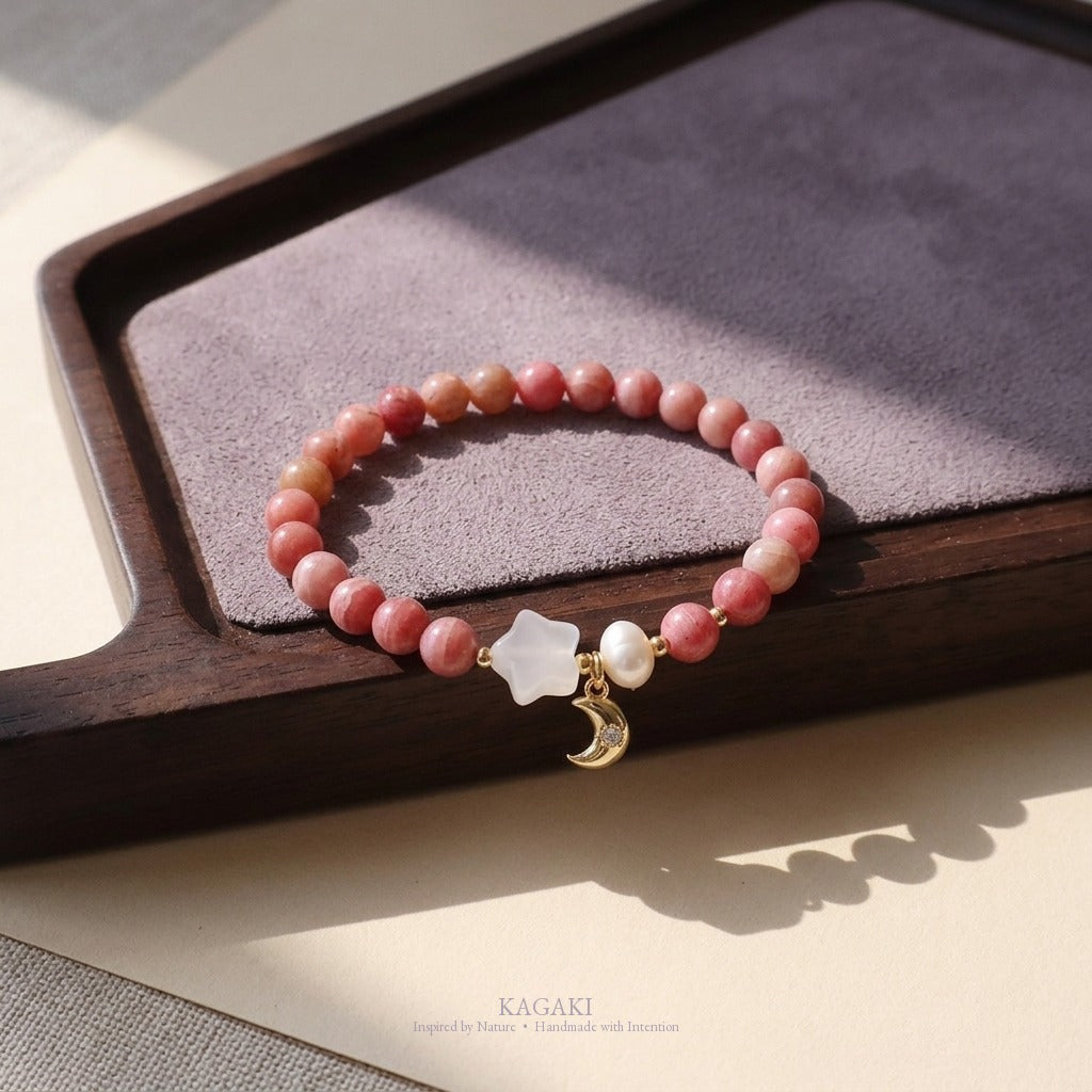 KAGAKI rhodochrosite bracelet with white chalcedony star, freshwater pearl, and gold-tone crescent moon charm styled on a dark walnut tea tray, raking natural daylight.