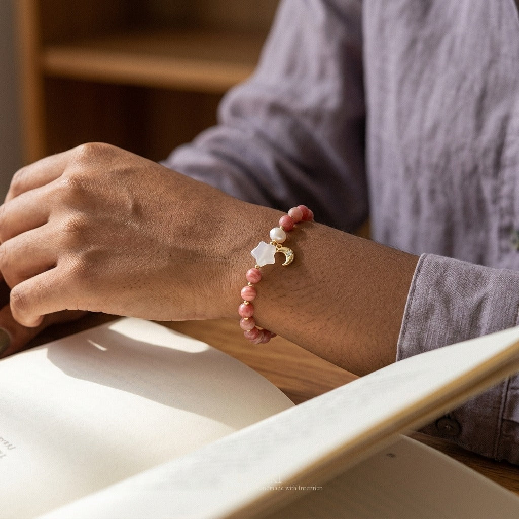 KAGAKI rhodochrosite bracelet with chalcedony star, pearl, and gold-tone crescent moon charm worn on a wrist turning the corner of an open paper book, side window light, editorial campaign styling.