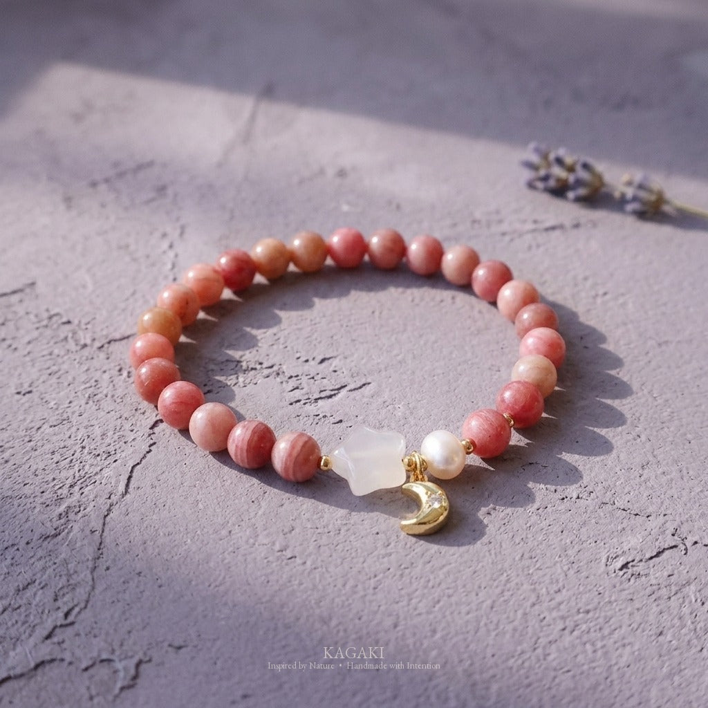 KAGAKI rhodochrosite bracelet with chalcedony star, pearl, and gold-tone crescent moon charm styled on weathered lavender-gray plaster with a dried lavender sprig.