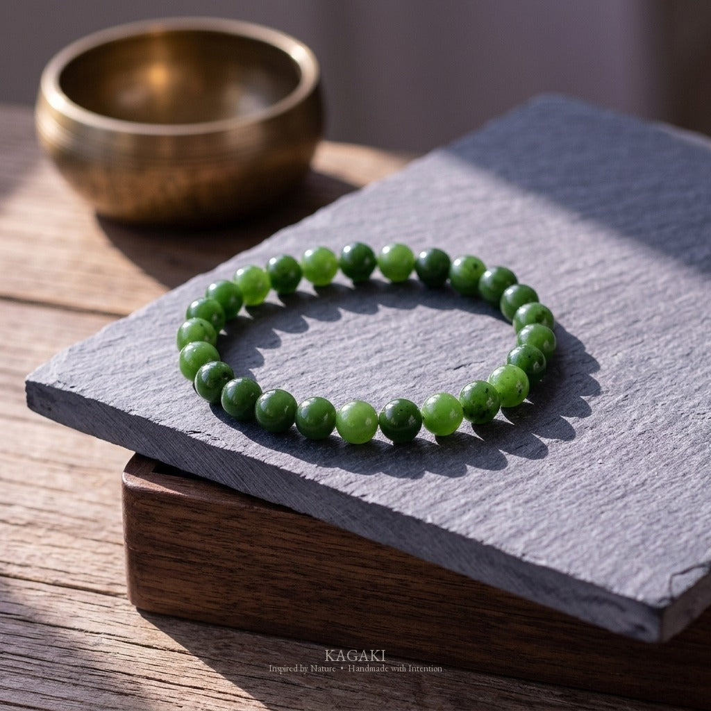 KAGAKI spinach-green jade bracelet styled on a slate slab with antique brass singing bowl and dark walnut box, raking afternoon daylight — quiet meditation altar editorial.