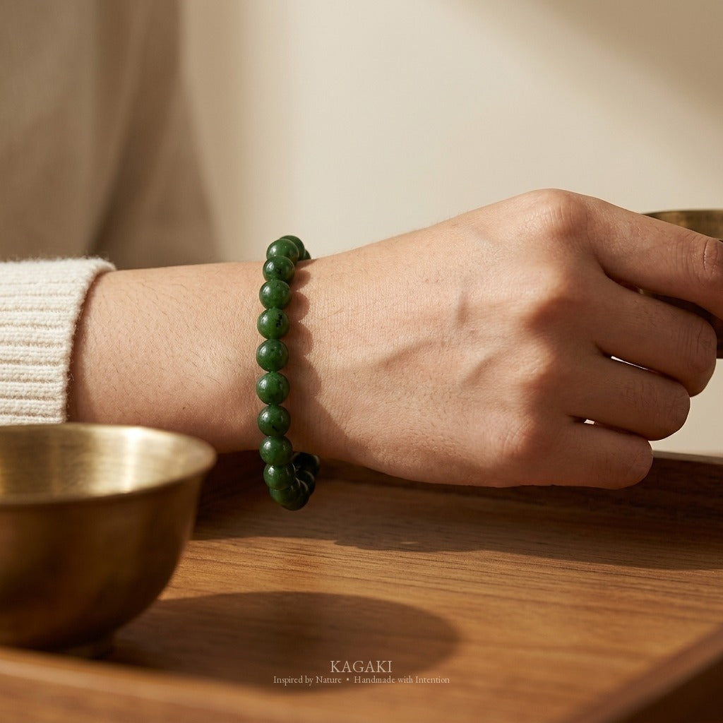 KAGAKI spinach-green jade bracelet worn on a wrist resting on a teak tray with a small antique brass tea bowl, fashion-editorial natural light.